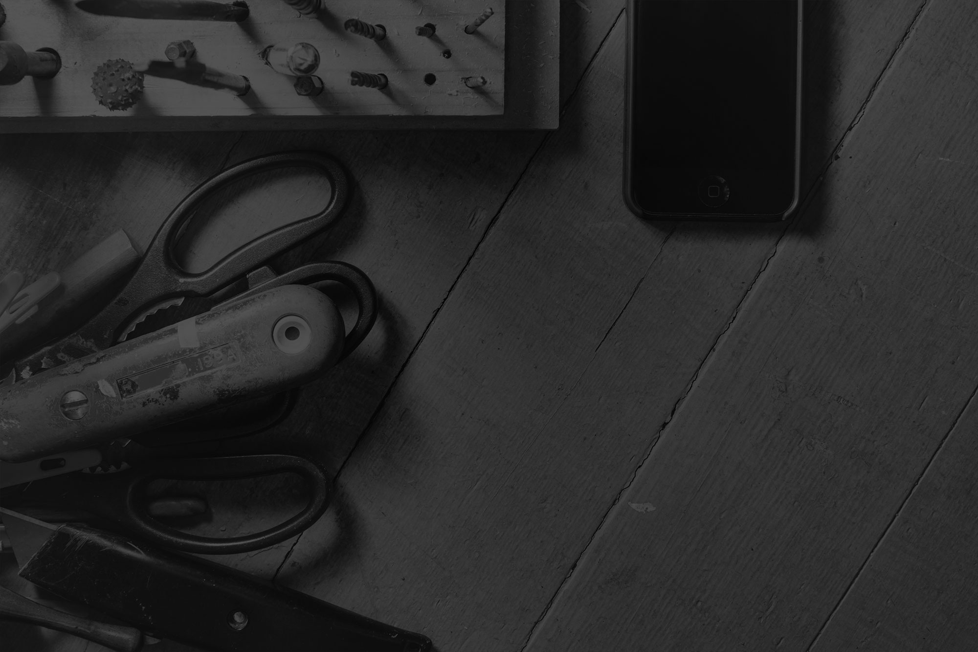 Close up black and white image of black iphone sitting on wooden work bench amongst old hand tools