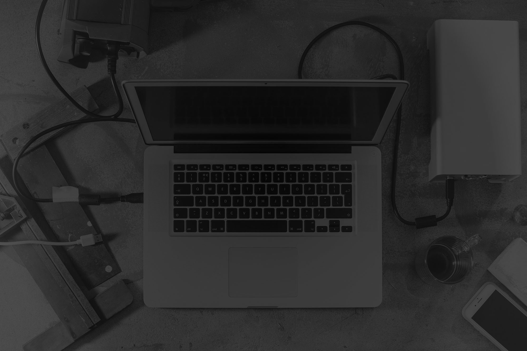 Black and white image of laptop, hard drive, iphone and coffee cup sitting on work bench amongst old hand tools